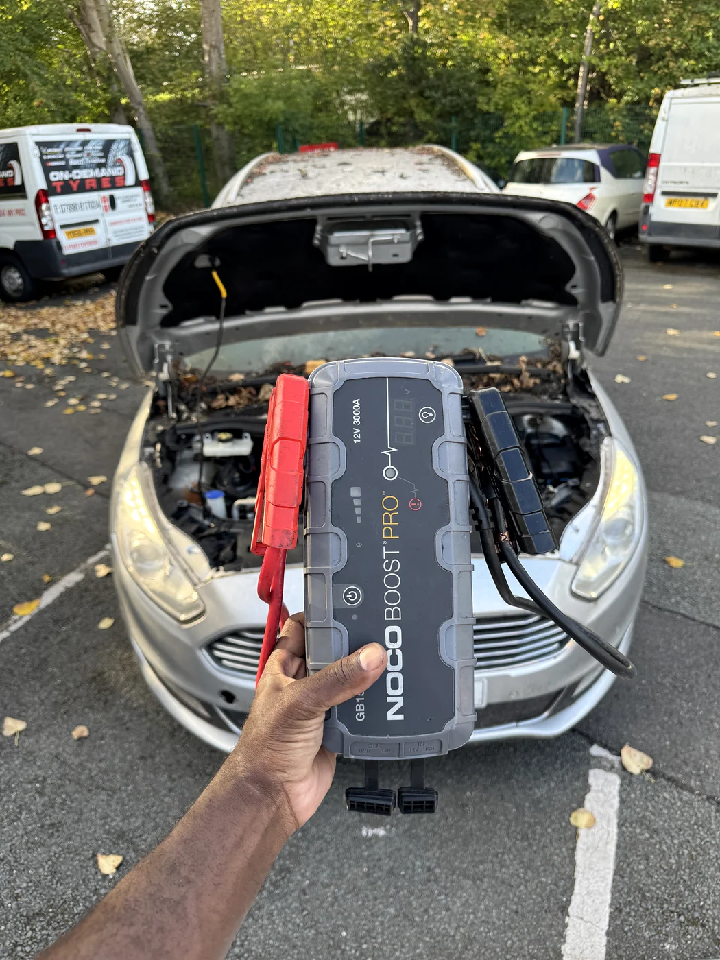 Portable NOCO super booster pack held in front of a car with its bonnet open