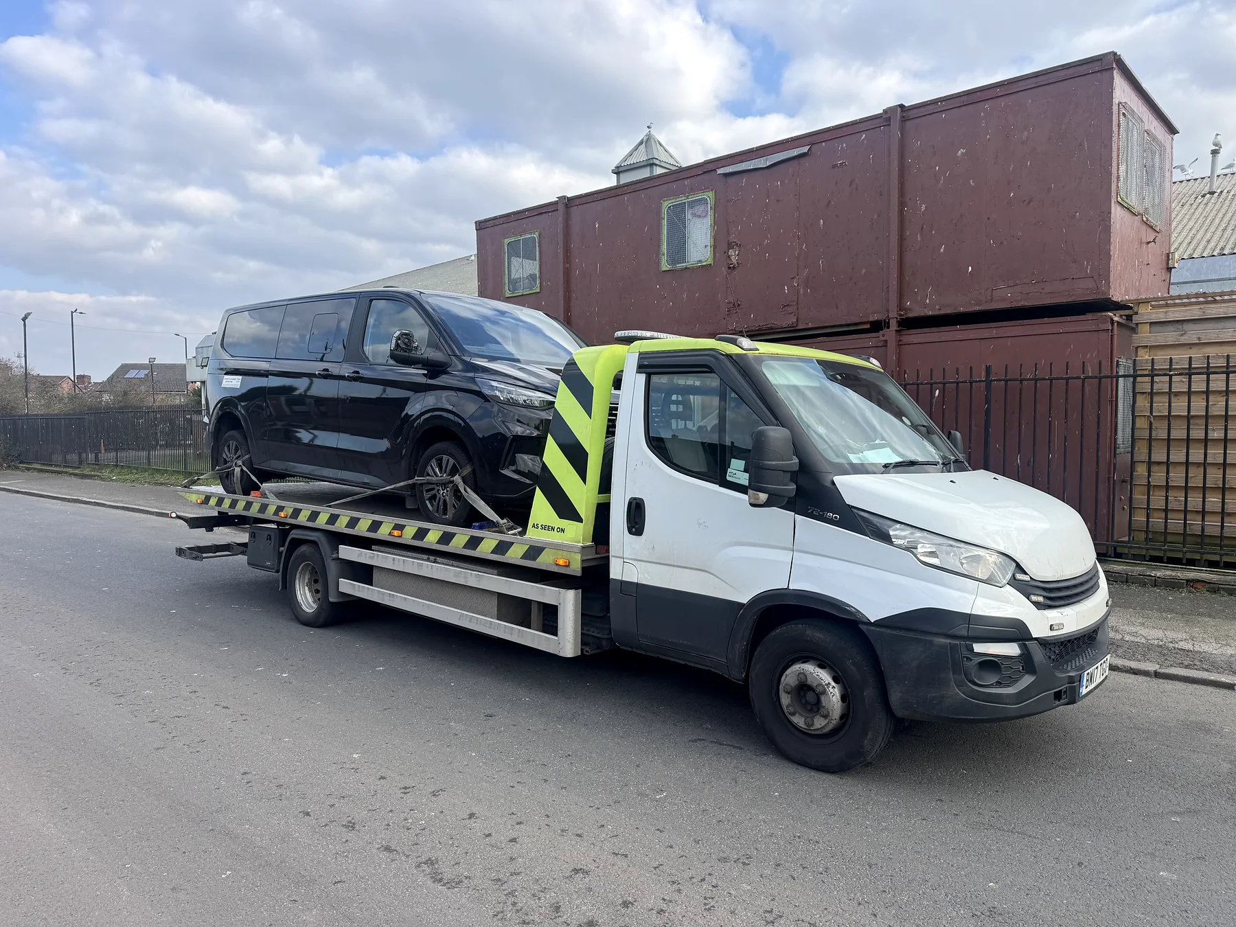 White Iveco carrying a dark van outside a storage yard